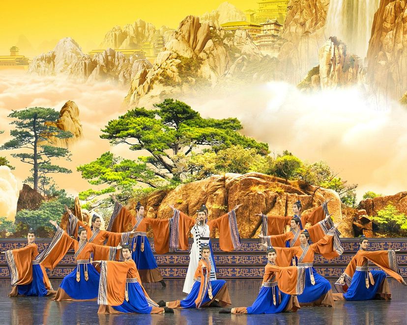 山水の背景幕の前で、白い衣の人物を囲み、橙と青の衣装の舞踊家たちが舞う物語の場面